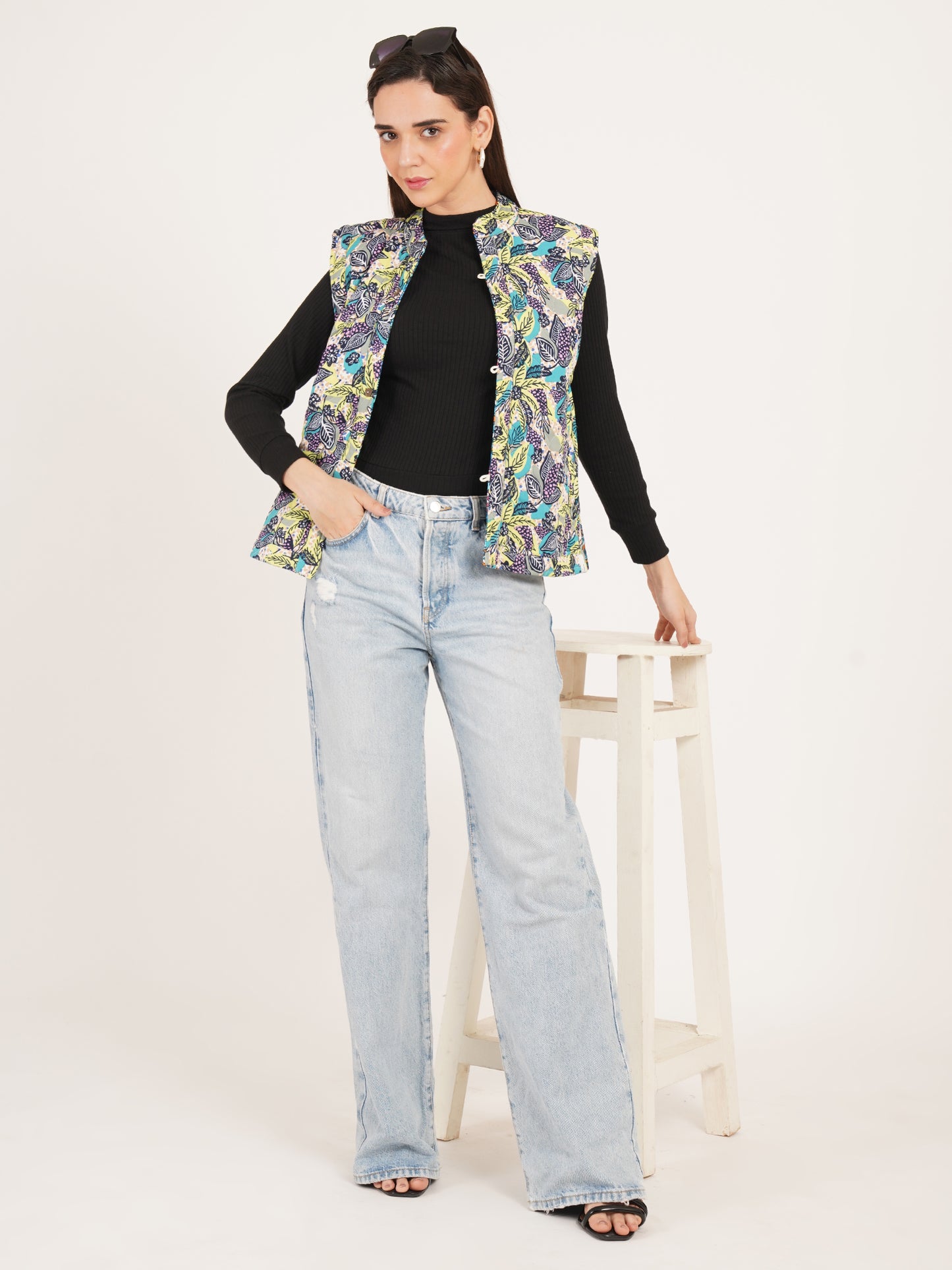 Woman sleeveless jackets wearing a floral vest over a black top and light blue jeans on a white background